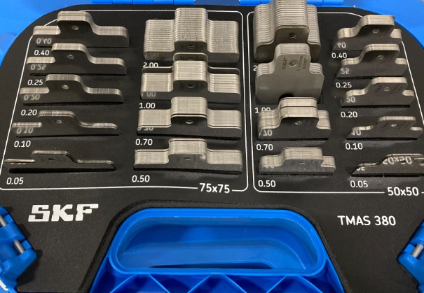 Wszechstronność i precyzja w jednej walizce. SKF TMAS 380 dla profesjonalistów. Obraz jest profesjonalną i kompletną prezentacją produktu, ukazującą zestaw SKF TMAS 380 jako wszechstronne i niezbędne narzędzie dla każdego profesjonalnego działu utrzymania ruchu. Kompozycja jest czysta, zorganizowana i techniczna. Dominują w niej lśniące metaliczne odcienie stali nierdzewnej podkładek, które kontrastują z ciemną, wytrzymałą walizką. Całość emanuje precyzją, wszechstronnością i gotowością do działania. Pierwszy plan – Kompletne rozwiązanie do osiowania: W centrum uwagi znajduje się solidna, otwarta walizka transportowa, która w pełni eksponuje bogate i uporządkowane wnętrze zestawu. Kluczowe elementy to: Wytrzymała walizka: Ciemna, przemysłowa walizka z wyraźnie widocznym białym logo "SKF" oraz oznaczeniem modelu "TMAS 380". Jej otwarta forma zaprasza do zapoznania się z imponującą zawartością. Bogaty zestaw podkładek: Wnętrze walizki, wyłożone specjalnie wyprofilowaną pianką lub przegródkami, zawiera imponującą liczbę 380 podkładek precyzyjnych w dwóch najpopularniejszych rozmiarach: 50 x 50 mm: Do mniejszych maszyn i precyzyjnych regulacji. 75 x 75 mm: Uniwersalny rozmiar do większości standardowych silników i pomp. Perfekcyjna organizacja: Każdy rozmiar podkładki ma swoją dedykowaną, wyraźnie oznaczoną sekcję, a w niej poszczególne stosy są uporządkowane według dziewięciu różnych grubości (od 0,05 mm do 2,00 mm). Taki układ wizualnie komunikuje łatwość i szybkość doboru odpowiedniej podkładki w każdych warunkach. Najwyższa jakość wykonania: Podkładki, wykonane z wysokogatunkowej stali nierdzewnej, lśnią w studyjnym oświetleniu. Na każdej z nich widoczne jest precyzyjne, laserowo grawerowane oznaczenie grubości i wymiaru, co eliminuje pomyłki. Charakterystyczne wycięcie ułatwiające montaż jest dobrze widoczne na każdym stosie. Tło i branding: Tło jest neutralne (np. białe lub jasnoszare), aby nie odwracać uwagi od bogactwa zestawu. Główne elementy brandingowe to logo SKF na walizce oraz oznaczenia na samych podkładkach, co jest gwarancją autentyczności i najwyższej jakości. Przesłanie i hasło reklamowe (optymalizacja SEO): Obraz niesie jasny komunikat o wszechstronności, precyzji i profesjonalizmie. Skierowany jest do techników utrzymania ruchu, firm serwisowych i inżynierów, którzy potrzebują jednego, kompletnego zestawu do osiowania maszyn o różnych gabarytach, pokrywającego większość standardowych zastosowań przemysłowych. Całość może być wzmocniona hasłem, które zawiera kluczowe frazy: "Jeden zestaw, pełna wszechstronność. Podkładki do osiowania SKF TMAS 380." "Wszechstronność i precyzja w jednej walizce. SKF TMAS 380 dla profesjonalistów." "Twoje kompletne rozwiązanie do osiowania maszyn – Zestaw SKF TMAS 380."